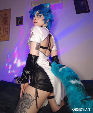 A person posing in a white and black cat-themed costume with a bright blue wig, cat ears, and a fluffy blue tail in a bedroom setting with purple ambient lighting.