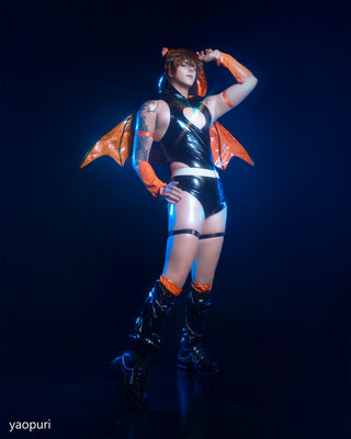 A person modeling a glossy black and orange fantasy-inspired costume against a dark blue studio background. The outfit includes a shiny black crop top and shorts with orange bat-like wing pieces, orange arm sleeves, thigh garters, and black knee-high boots.