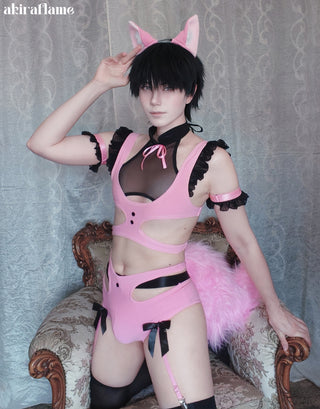 A person wearing a pink and black feline-inspired lingerie costume with pink cat ears, a fluffy pink tail, and thigh garters, posed on an ornate vintage armchair against a patterned gray curtain backdrop.